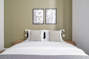 Ein Bett mit weißer Bettwäsche, zwei Kissen und einer grauen Decke. Über dem Bett hängen zwei gerahmte abstrakte Kunstwerke an der Wand.