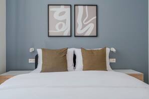 A bed with white bedding and two brown pillows, a nightstand with a white lamp, and two framed abstract art pieces on the wall. - Blueground | Holešovice, city view, nr Vltava (PRG-53) (Prague)