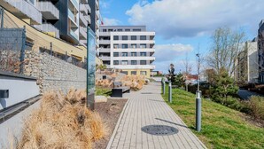 En gång leder fram till en modern byggnad med flera våningar, en stenmur och ett grönområde med bänkar.