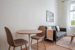 Ein Wohnzimmer mit rundem Tisch, zwei Stühlen, einer Couch und einem Fenster mit Vorhängen.