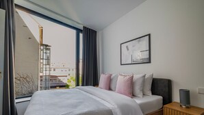 Quarto com cama, mesa de cabeceira, abajur, janela com cortinas e um quadro na parede.