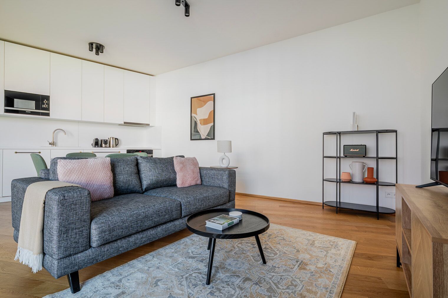 A modern living room with a grey sofa, a coffee table, a bookshelf, and a kitchen area in the background.