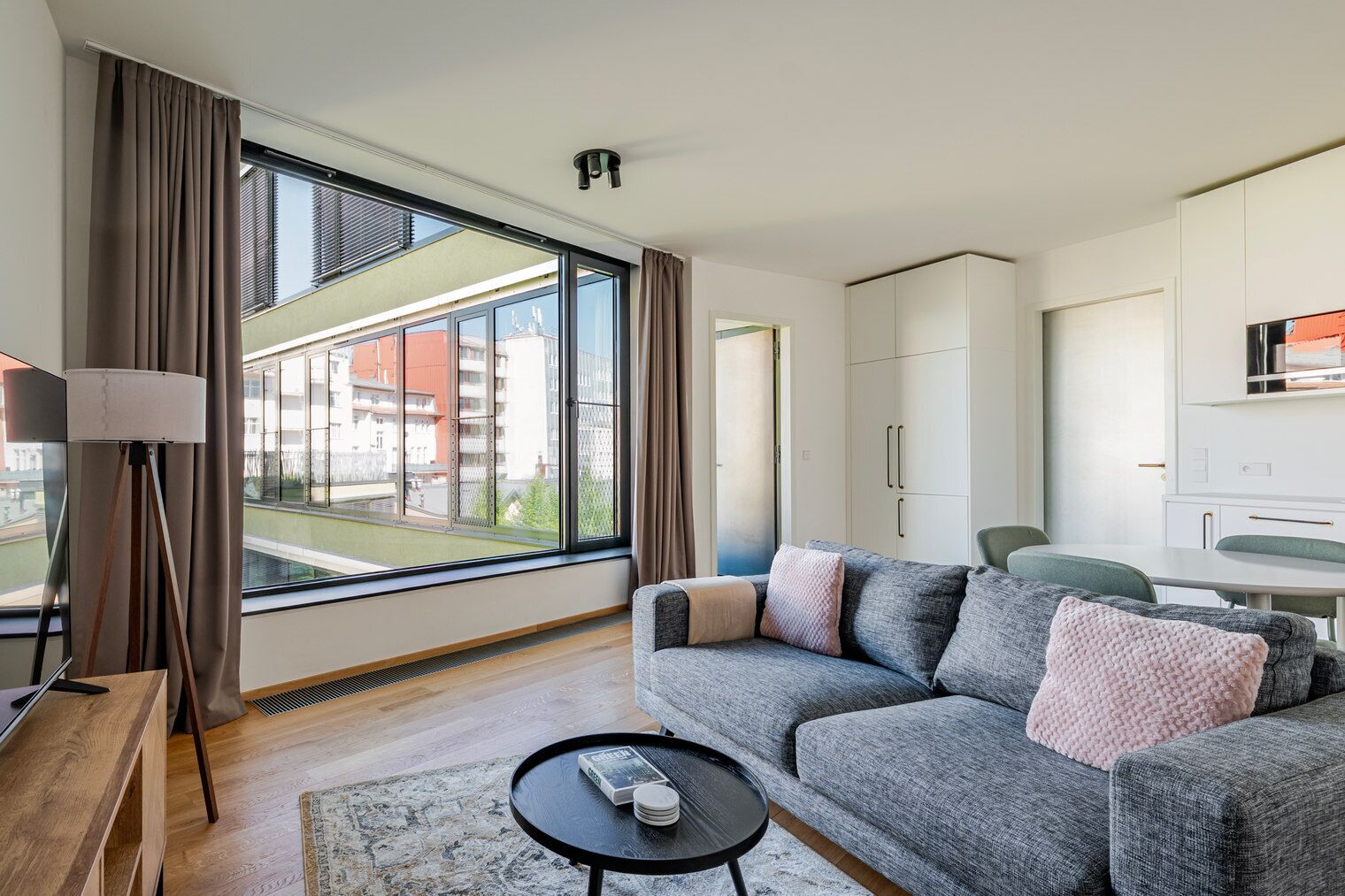 Ett modernt vardagsrum med en soffa, ett soffbord och en lampa. Det finns ett stort fönster med gardiner, utsikt över byggnader utanför och en liten köksdel.