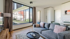 Uma sala de estar moderna com um sofá, uma mesa de centro e uma luminária. Há uma janela grande com cortinas, vista de edifícios e um espaço com cozinha compacta.