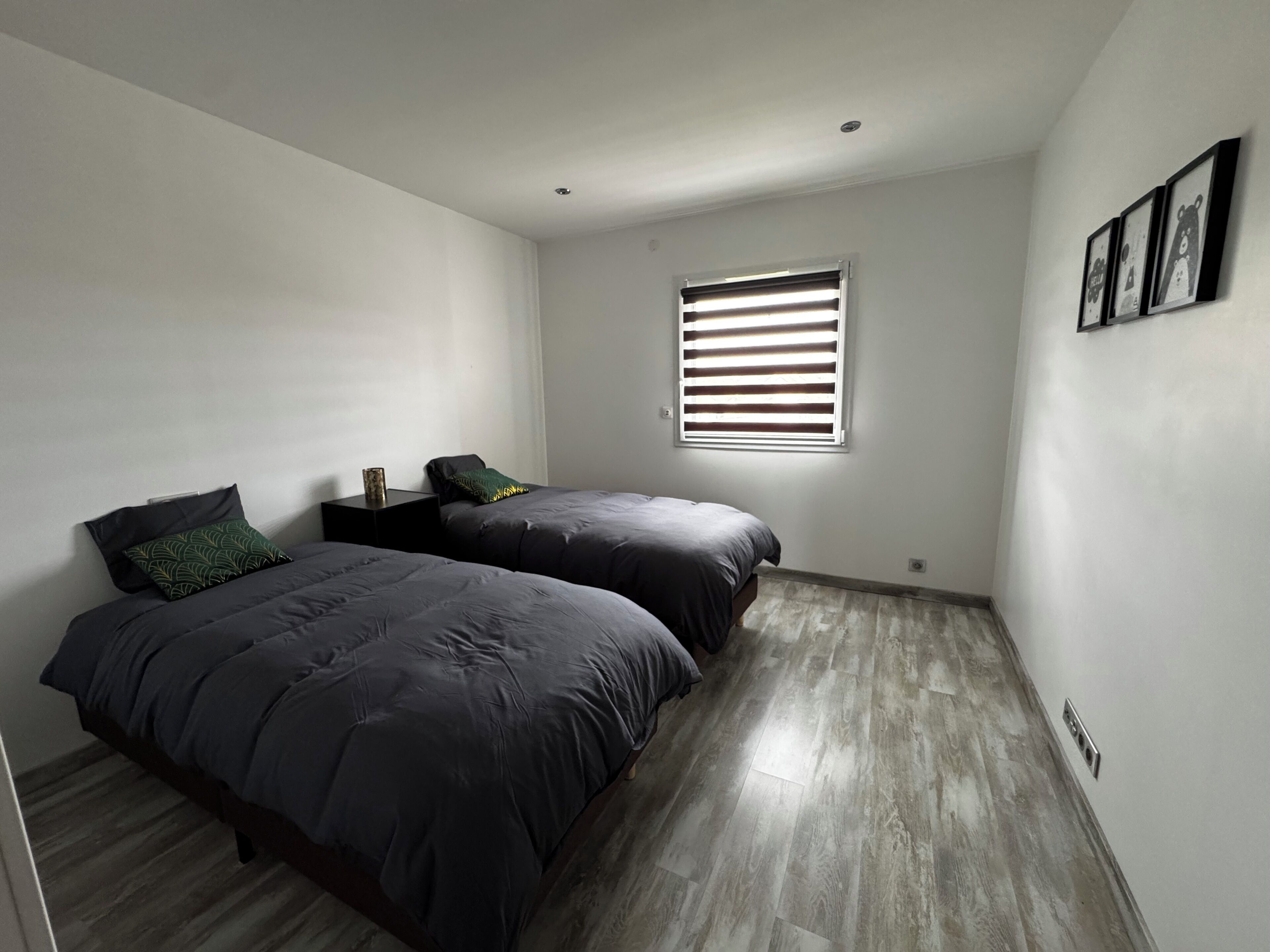A bedroom with two beds, a window with blinds, a nightstand, and a wall-mounted light fixture.
