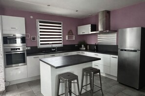 Une cuisine moderne avec des meubles blancs, des appareils en acier inoxydable, un îlot central doté de tabourets et une fenêtre avec des stores.