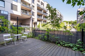A wooden deck with two white chairs and a table, surrounded by a black metal fence and greenery. - Blueground | Smíchov, rooftop, nr Vltava River (PRG-106) (Prague)