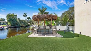 Eine Terrasse mit einem strohgedeckten Pavillon, einem Esstisch mit Stühlen, einem Pool und umgebender Bepflanzung.