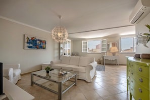 Ein Wohnzimmer mit einem weißen Sofa, einem Glastisch, einem Gemälde und einer Kronleuchter.