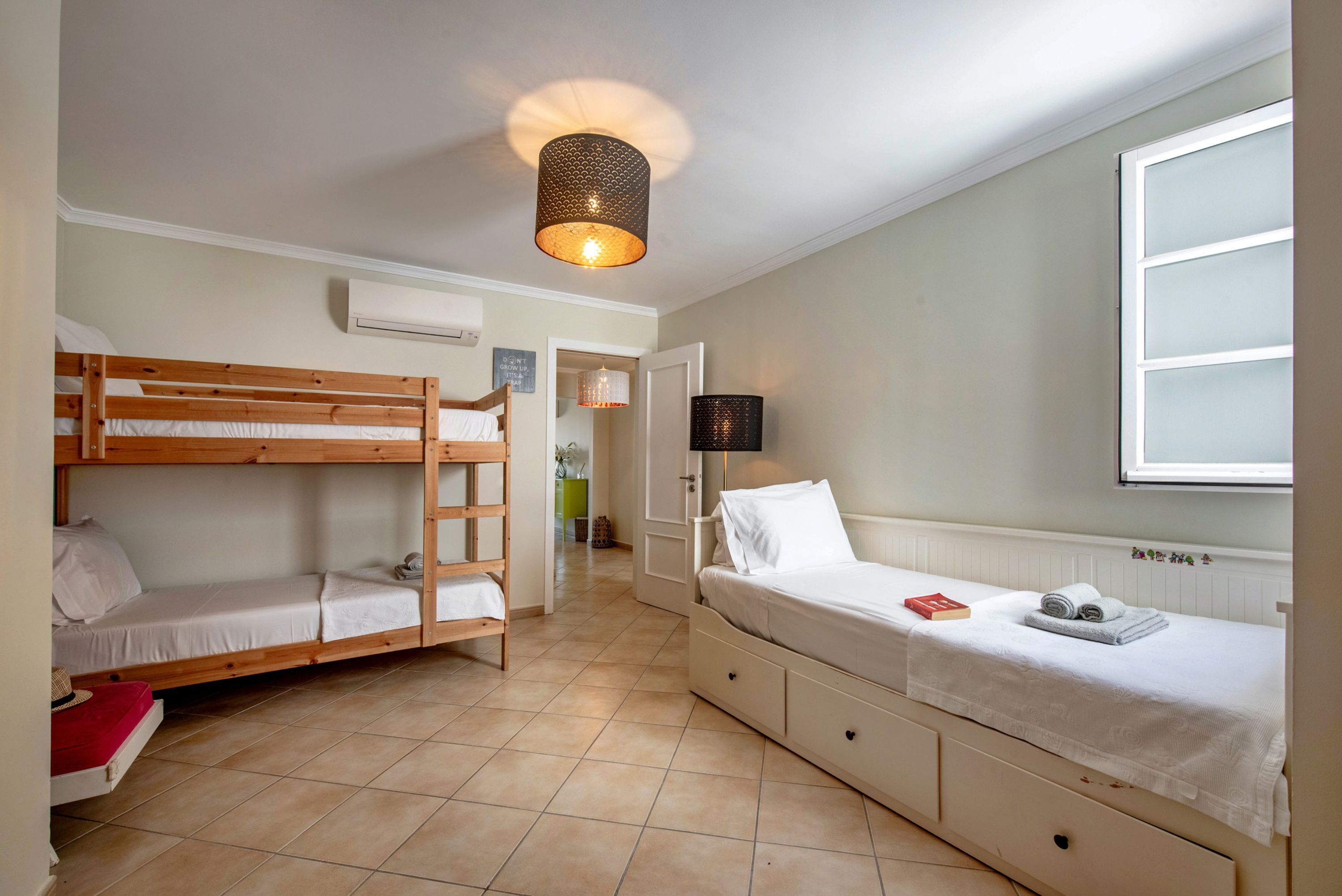 Une chambre avec un lit superposé, un lit simple, une lampe, une fenêtre et un appareil de climatisation.