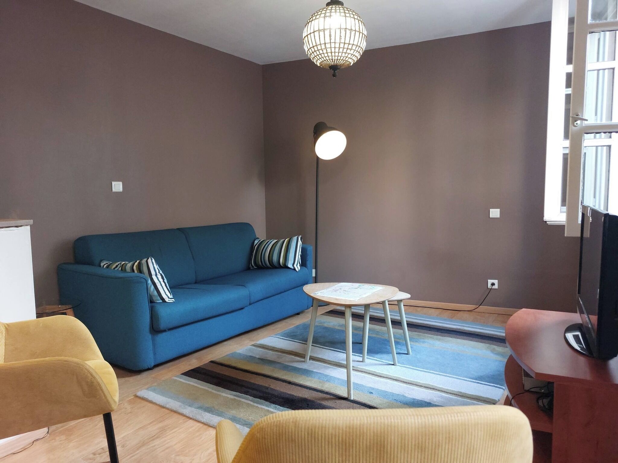 A living room with a blue sofa, a yellow armchair, a small round table, a floor lamp, and a TV on a wooden stand.