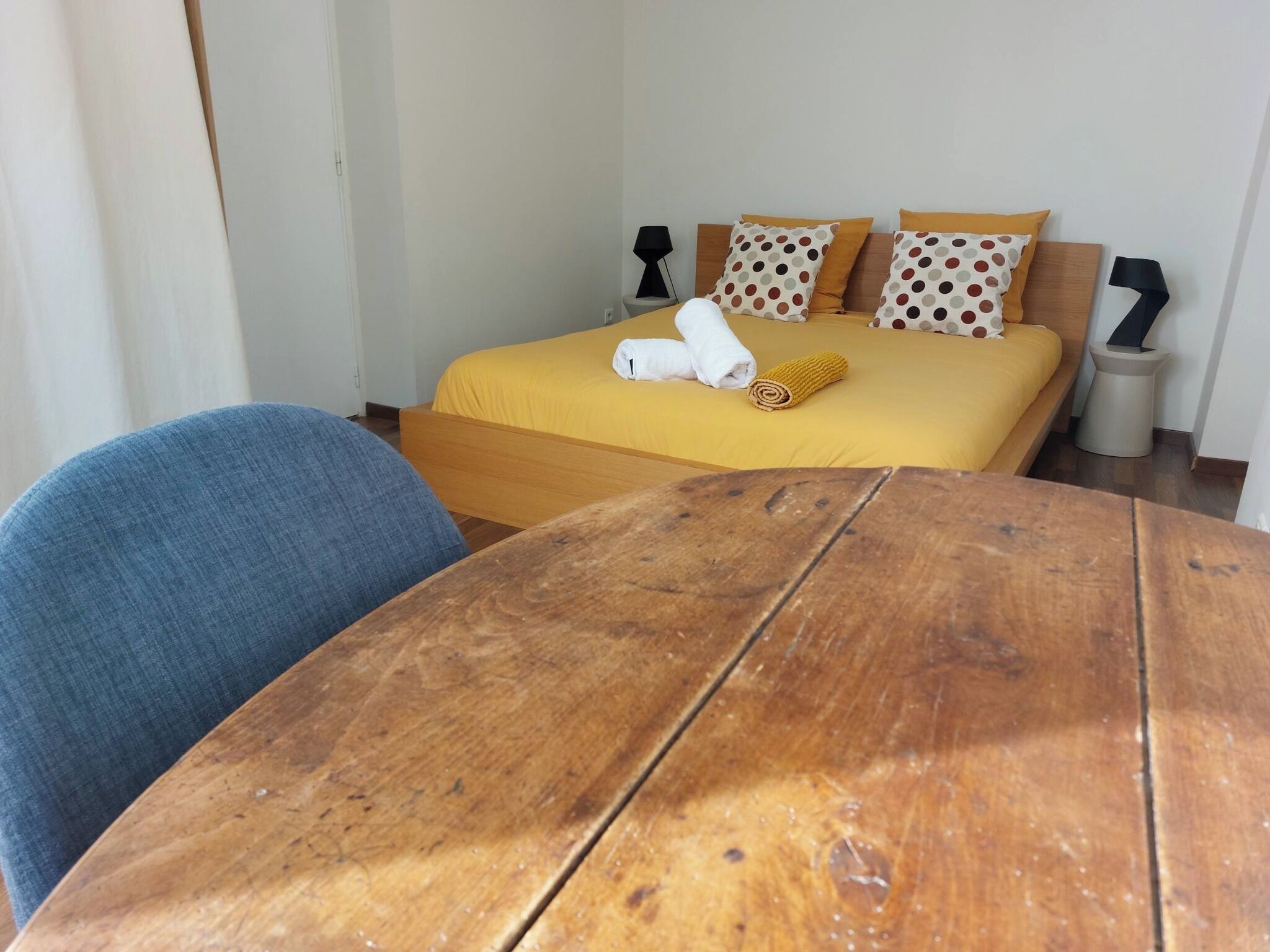 Une chambre d’hôtel avec une table en bois, une chaise bleue, un lit avec une literie jaune et deux tables de chevet avec des lampes.