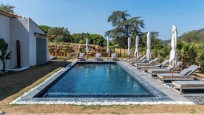 Une piscine rectangulaire, agrémentée de chaises longues et de parasols, entourée d’un bâtiment blanc et de verdure.