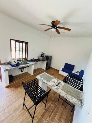 A compact kitchen with a window, a ceiling fan, and a dining area with two chairs. - Nacho Apartments (Puerto Vallarta)