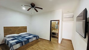 A bedroom with a bed, a ceiling fan, a wooden headboard, a bathroom with a mirror, and a TV mounted on the wall. - Nacho Apartments (Puerto Vallarta)