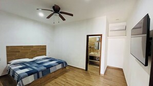 A bedroom with a bed, a ceiling fan, a wooden headboard, a bathroom with a mirror, and a TV mounted on the wall. - Nacho Apartments (Puerto Vallarta)