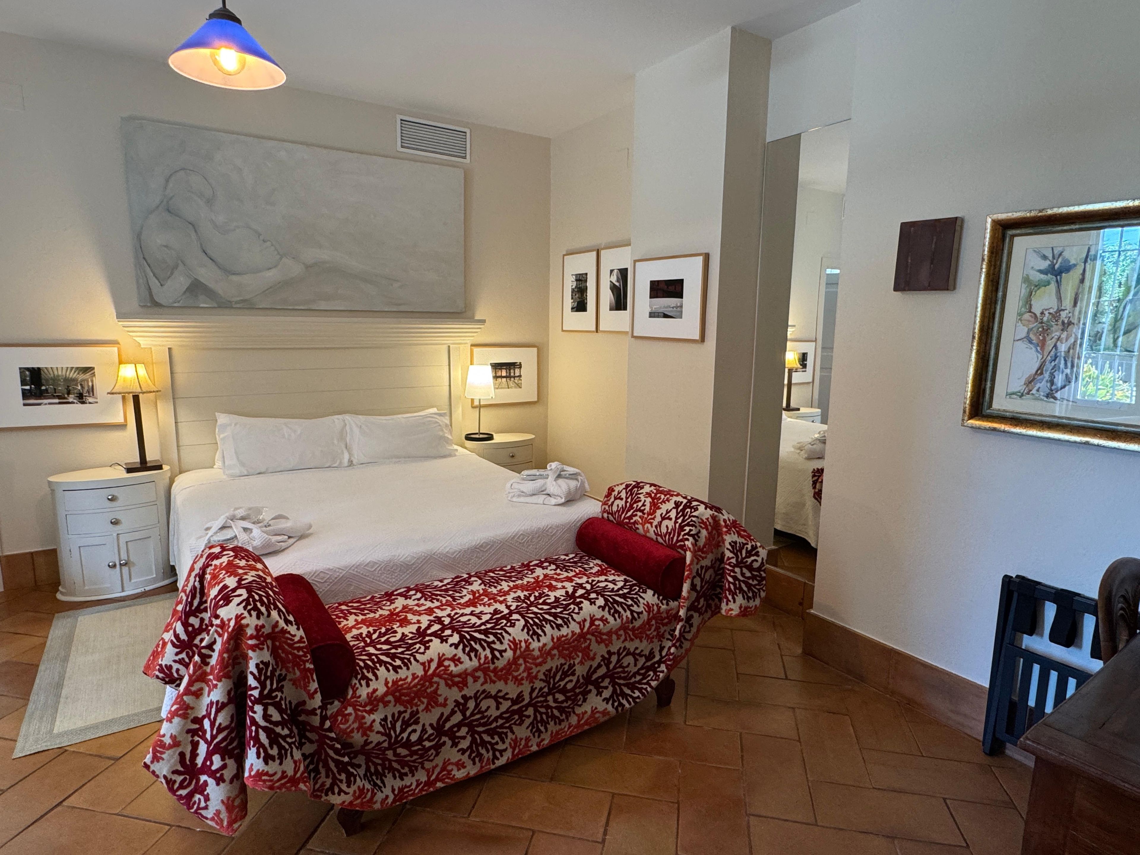 A hotel room with a bed, bedside lamps, a large painting, framed pictures, and a sofa.