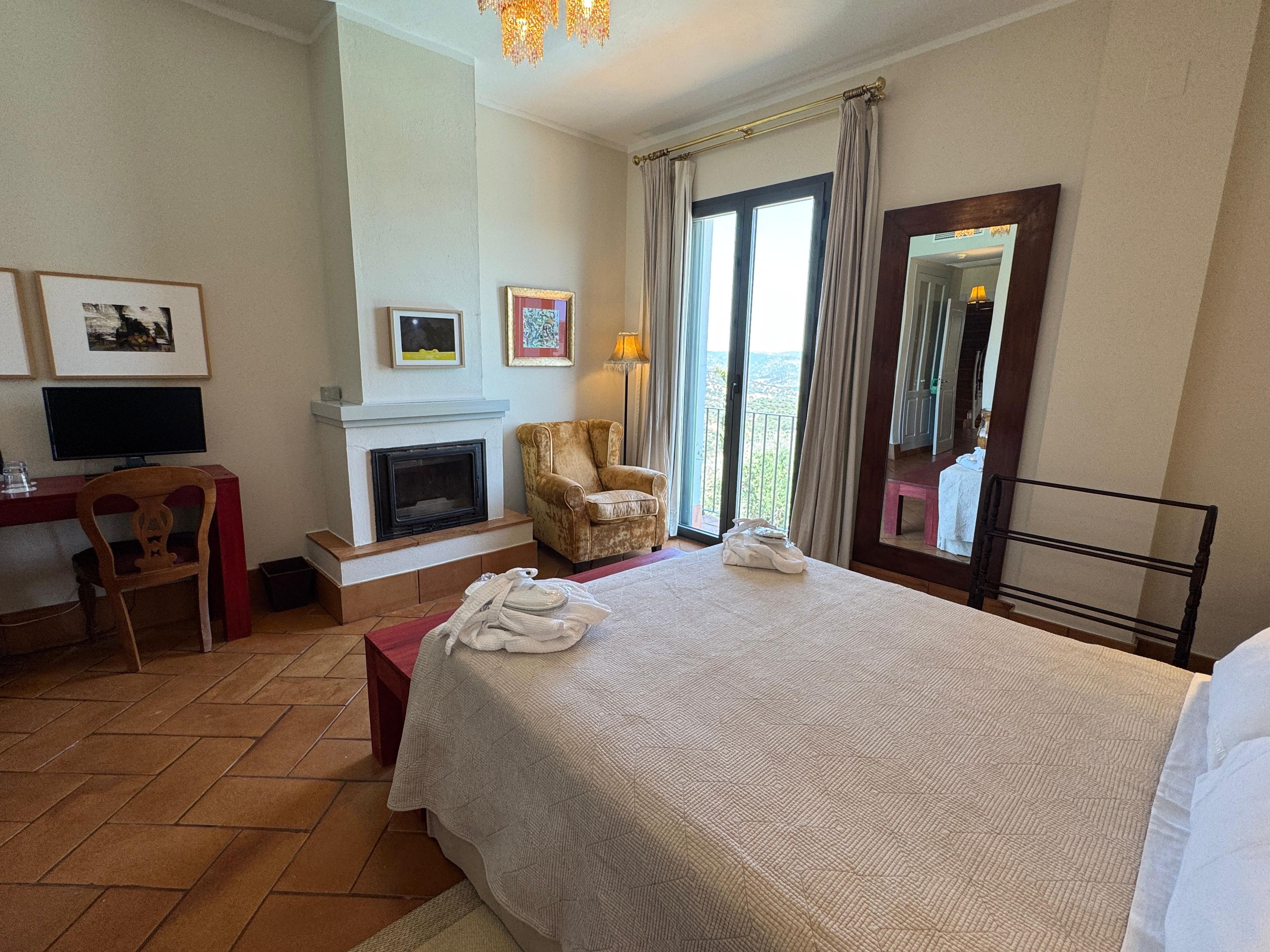 Habitación de hotel con cama, escritorio con computadora, chimenea, silla y vistas al balcón.