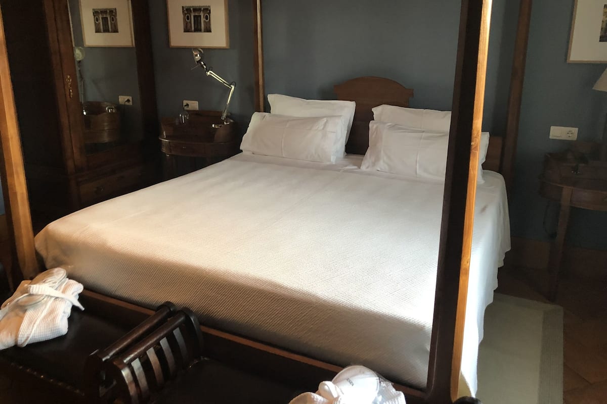 A four-poster bed with white linens, a wooden headboard, and a nightstand with a lamp.