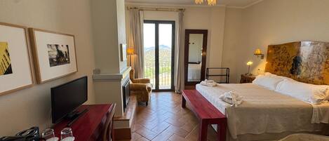 A hotel room with a bed, a desk with a TV, a chair, a bench, and a view of the outdoors.