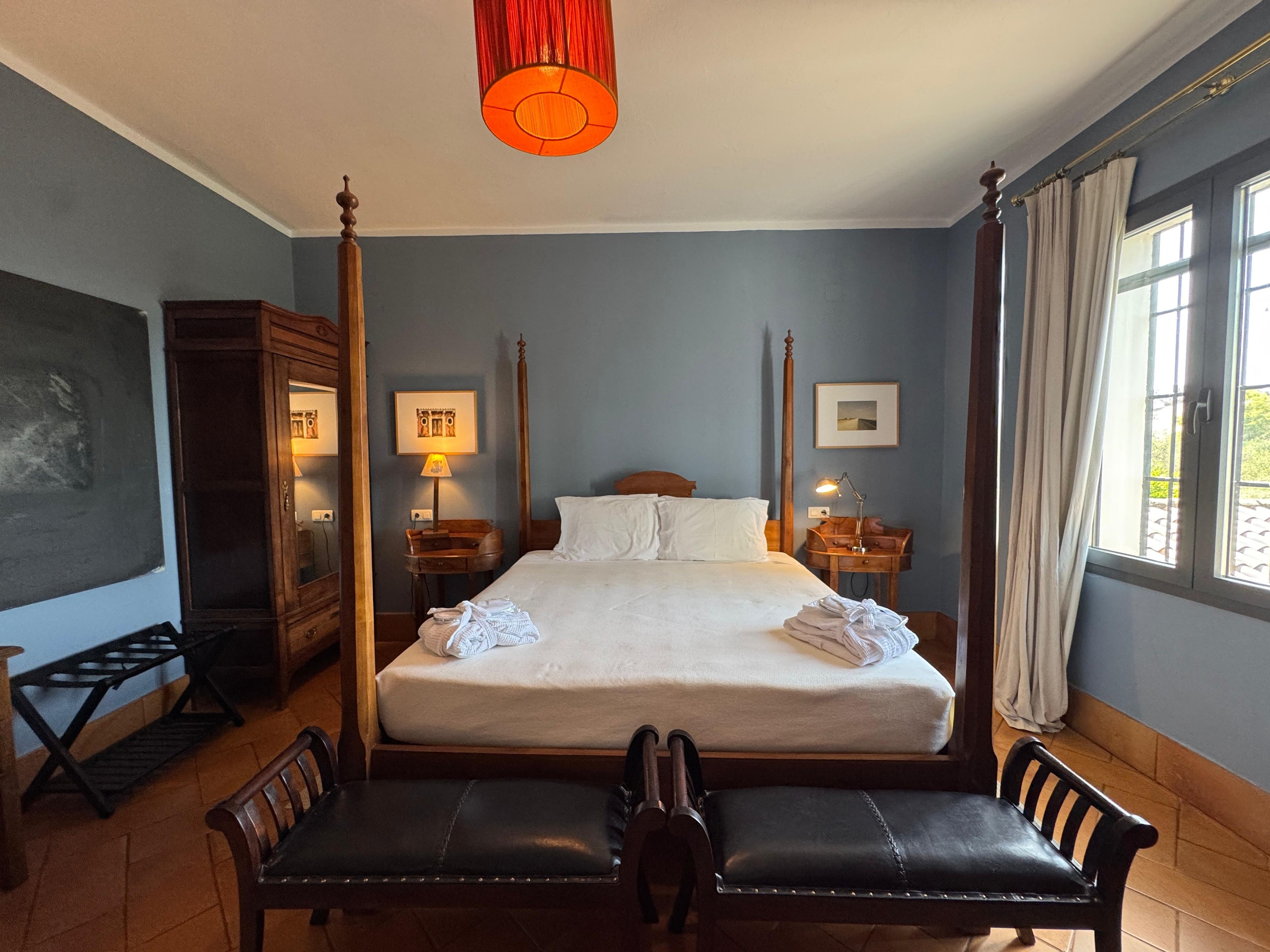 Habitación de hotel con cama con dosel, un escritorio con lámpara, una silla y un gran ventanal con cortinas.