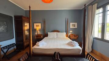 Habitación de hotel con cama con dosel, un escritorio con lámpara, una silla y un gran ventanal con cortinas.