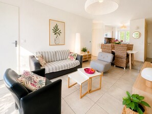 Ein modernes Wohnzimmer mit einer Couch, einer Essecke mit Tisch und Stühlen, einer Küche im Hintergrund und einem großen Fenster mit Grünpflanzen draußen.