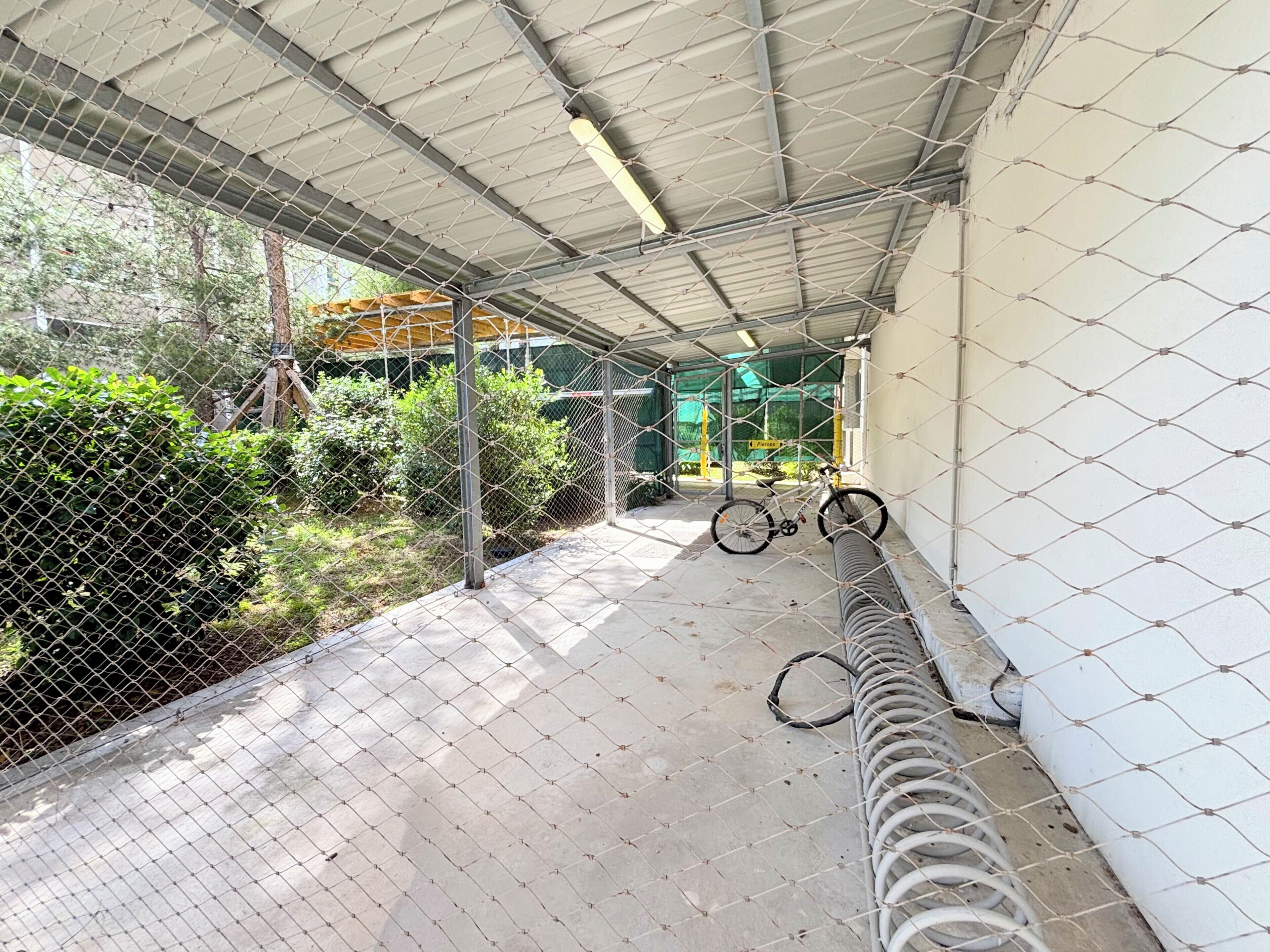 Un espacio al aire libre techado, con una cerca de malla metálica, una bicicleta y una manguera enrollada en el suelo.