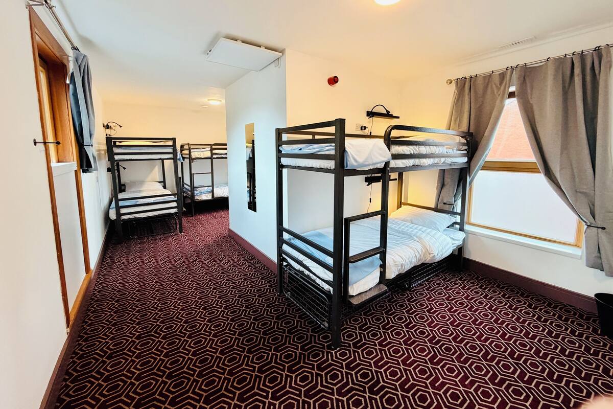 A dormitory room with bunk beds, a patterned carpet, a window with curtains, and a ceiling light.
