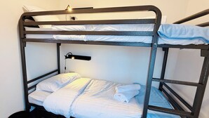A bunk bed with white bedding, a small black trash bin, and a wall-mounted light fixture.