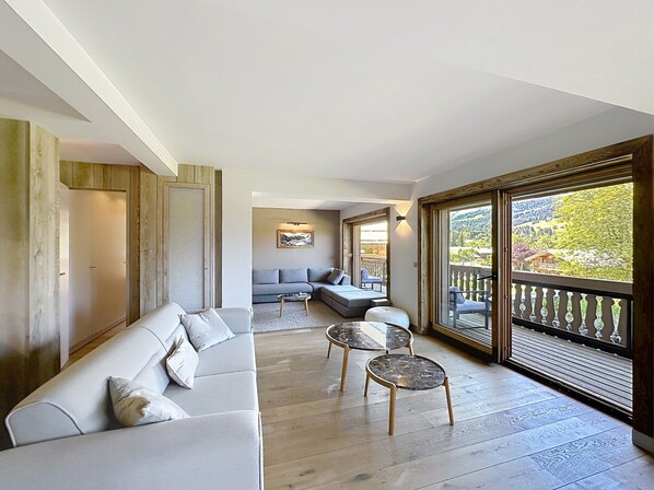 Een ruime zithoek met een bank, twee bijzettafels en een balkon met uitzicht op een bebost landschap.