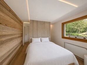 Ein Schlafzimmer mit Holzwand, einem Bett mit weißer Bettwäsche, einem Fenster mit Blick auf Grünflächen und Einbauleuchten an der Decke.