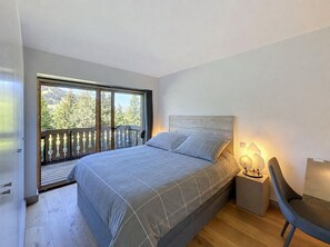 Een slaapkamer met een groot raam, een bed, een nachtkastje en een stoel.