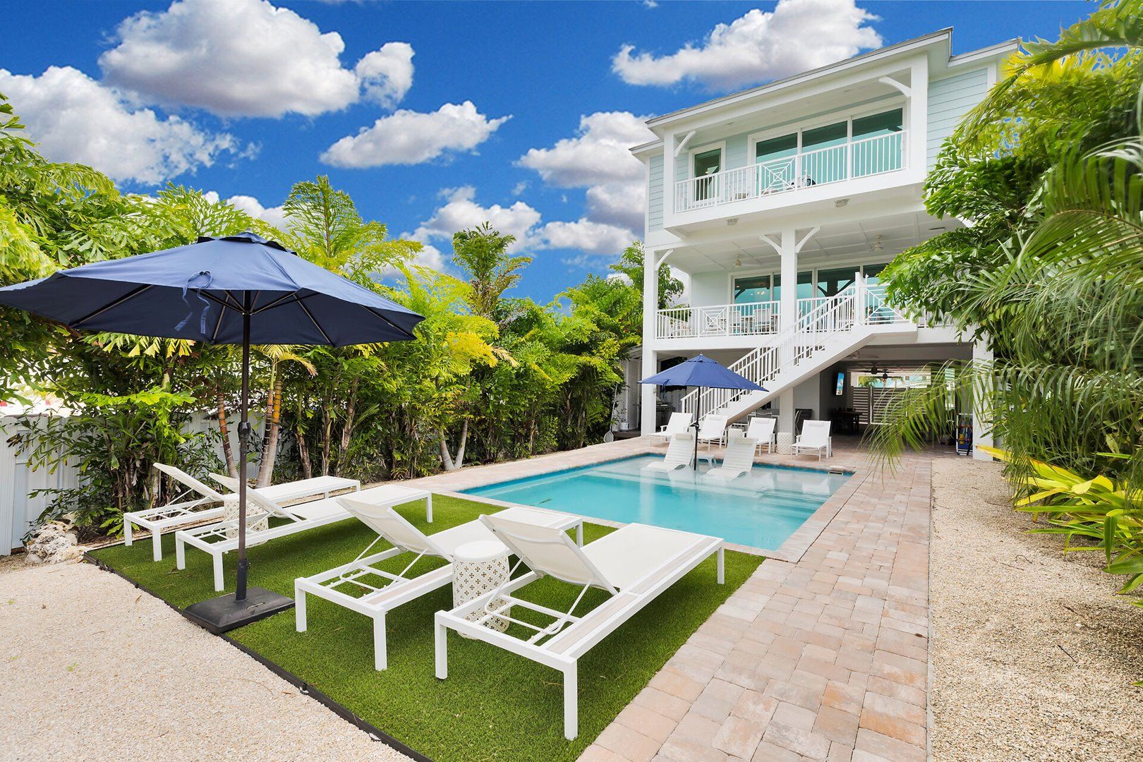 A pool area with lounge chairs and umbrellas, surrounded by lush greenery and a two-story building.
