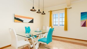 A dining area with a glass table, white and blue chairs, pendant lights, a painting, and a window with yellow curtains. - Desertas Vista Casa by LovelyStay (Gaula)