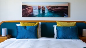 Een bed met witte beddengoed, twee blauwe kussens en een geel kussen, met daarboven een schilderij van een kustlandschap.