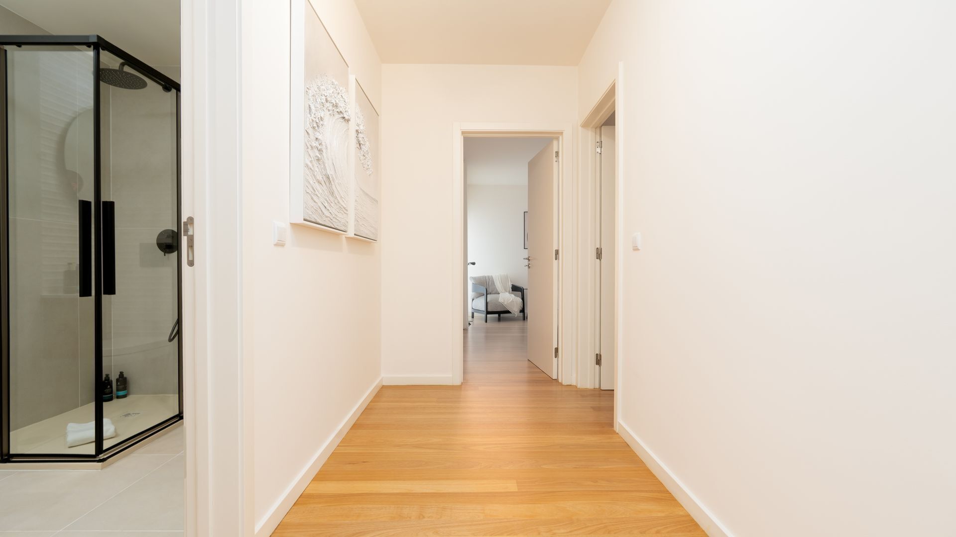 A hallway with a glass shower enclosure, a wooden floor, and a door leading to a room with a bed and a chair.