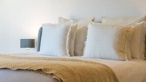 Ein Bett mit weißen Leinen und einer beige farbenen Decke, eine Nachttischlampe und eine schlichte Wand.