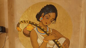 A bed with white and brown pillows, a wall with a mural of a person playing a stringed instrument, and a ceiling light. - Shades of Kashi with an artistic and cozy stays for couple and family. (varanasi)
