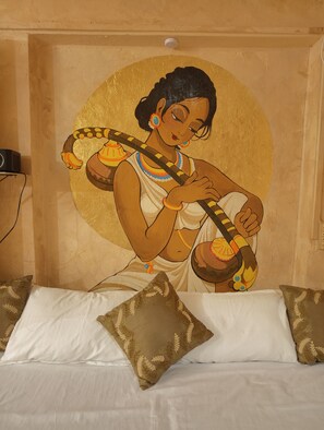 Un letto con cuscini bianchi e marroni, un muro con un affresco che raffigura una persona che suona uno strumento a corda e una lampada a soffitto.