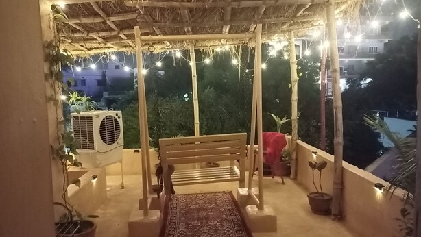 A balcony with a wooden bench, a patterned rug, potted plants, and string lights. - Shades of Kashi with an artistic and cozy stays for couple and family. (varanasi)