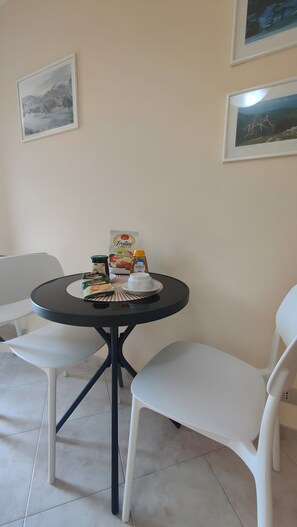 Un'area pranzo con un tavolo rotondo, due sedie bianche e un piccolo tavolo bianco. Alle pareti ci sono quadri incorniciati, una scatola di pasta Frollini, un vasetto di marmellata e una bottiglia di latte.