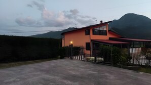 Uma casa de dois andares com telhado vermelho, um caminho de entrada pavimentado e uma montanha ao fundo.