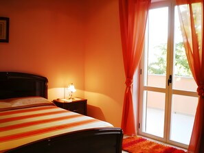 Una habitación con una cama, una mesita de noche con lámpara y un ventanal con cortinas.