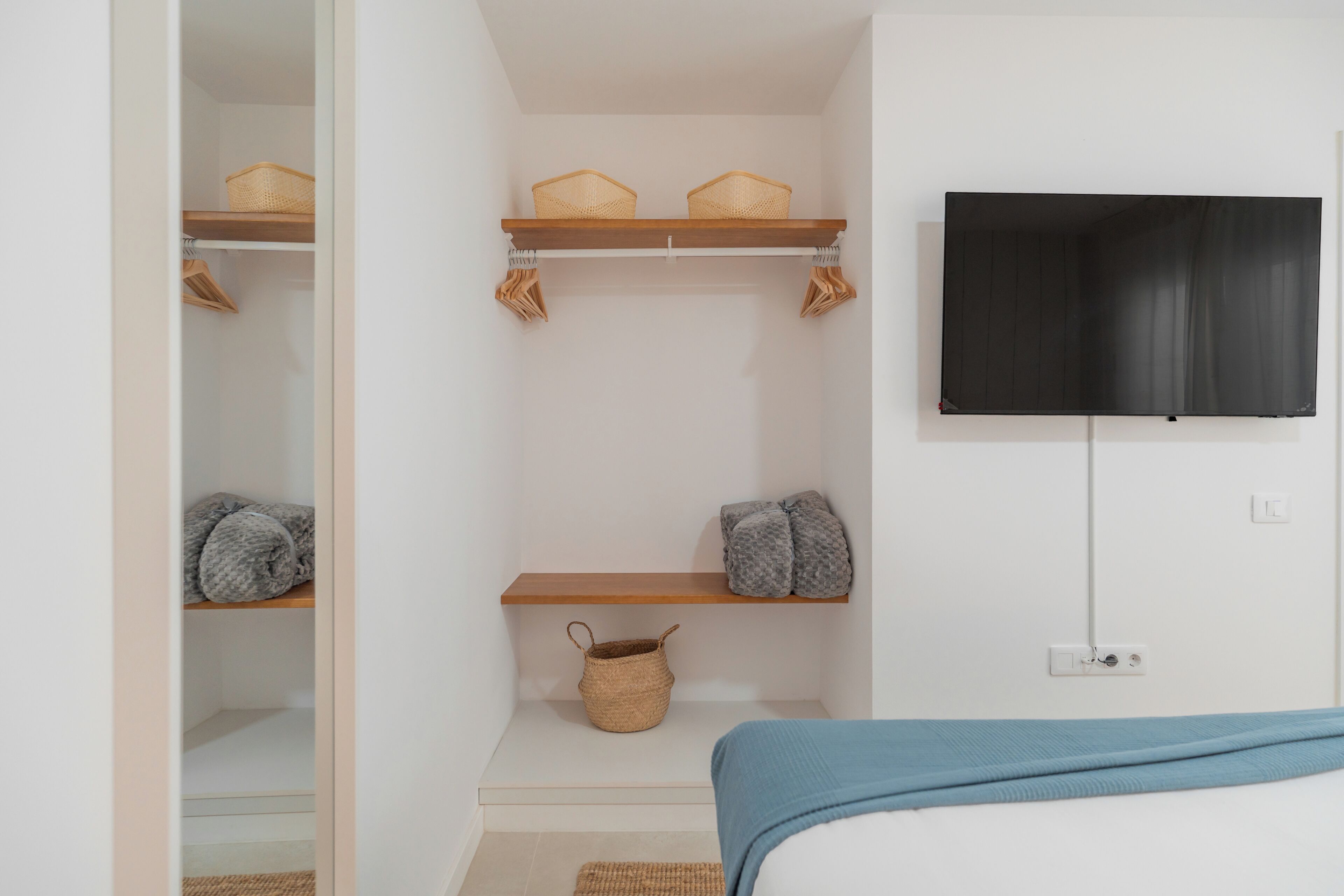 A bedroom with a bed, a TV mounted on the wall, and shelves with baskets and folded blankets.