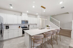 Une cuisine moderne avec des meubles blancs, un îlot central doté de tabourets de bar, des appareils en acier inoxydable et un escalier menant à l’étage supérieur.