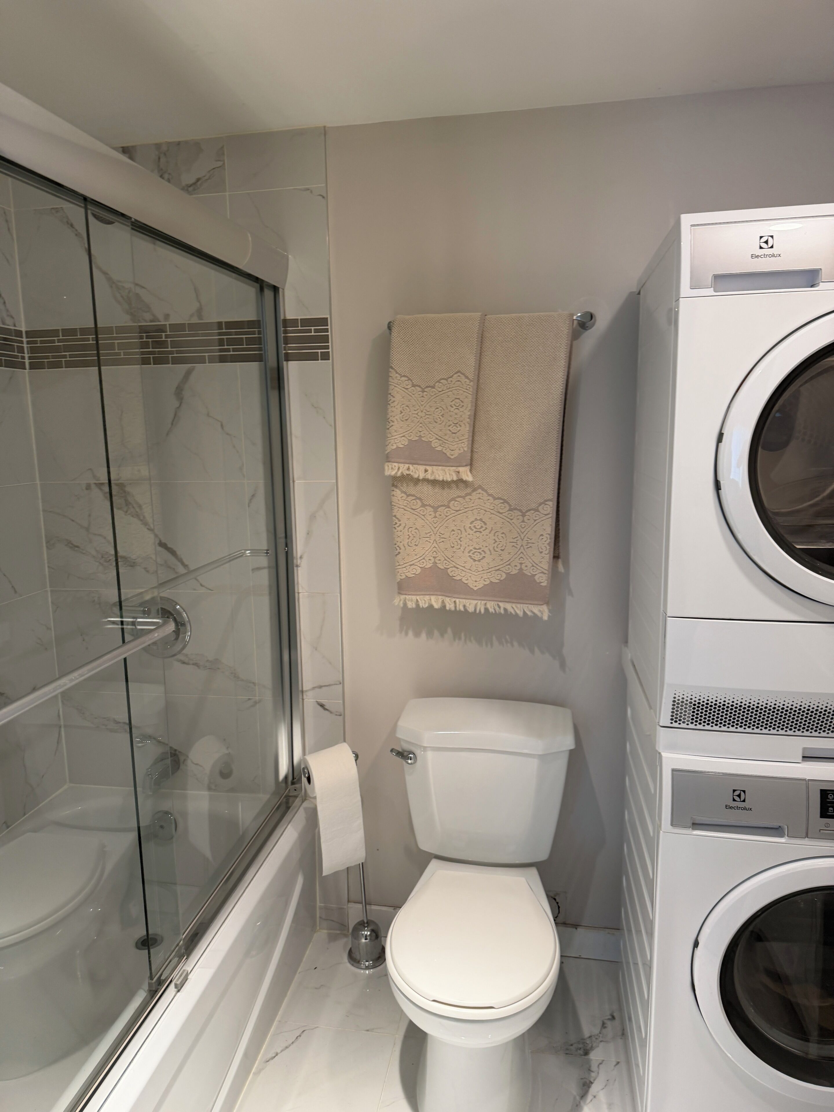 A bathroom with a glass-enclosed shower, a toilet, and a stacked washer and dryer unit.