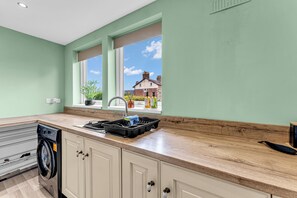 A kitchen with a washing machine, wooden countertop, and a window with a view of houses. - Haregate House | 3BR Leek | Peak District | Sleeps 5 (Leek)