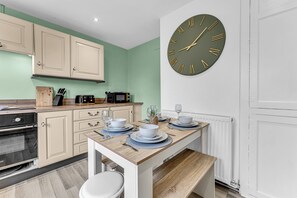 A modern kitchen with a wooden table set for dining, a large wall clock, and light green cabinets. - Haregate House | 3BR Leek | Peak District | Sleeps 5 (Leek)