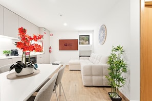 Ein modernes Wohnzimmer mit einem weißen Sofa, einem Esstisch mit Stühlen, einer Küchenzeile und einem großen Fenster.